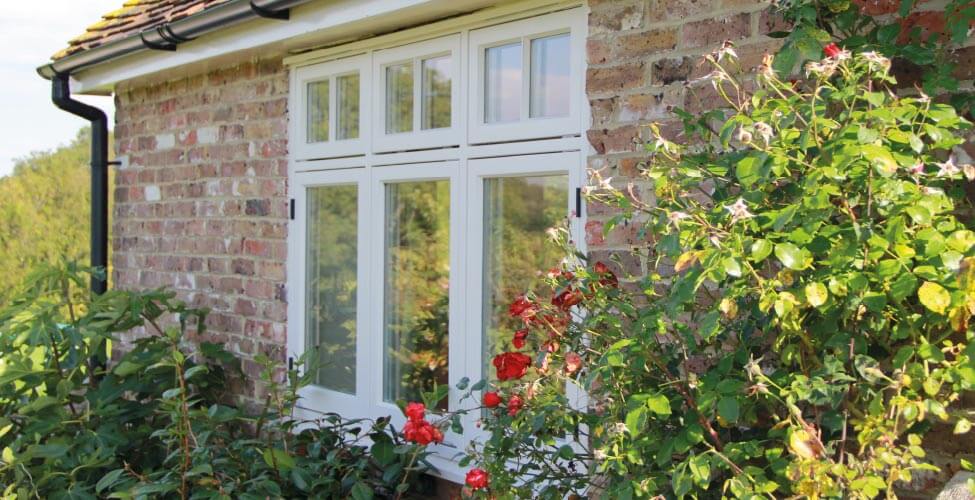 A white-framed timber window is set in a brick wall, surrounded by blooming red roses and green foliage in a sunny garden.