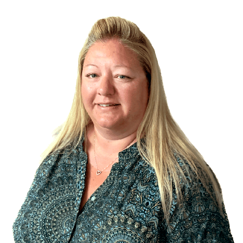 Nicola with long blonde hair is smiling gently, wearing a patterned teal blouse with a heart-shaped necklace, against a plain white background.